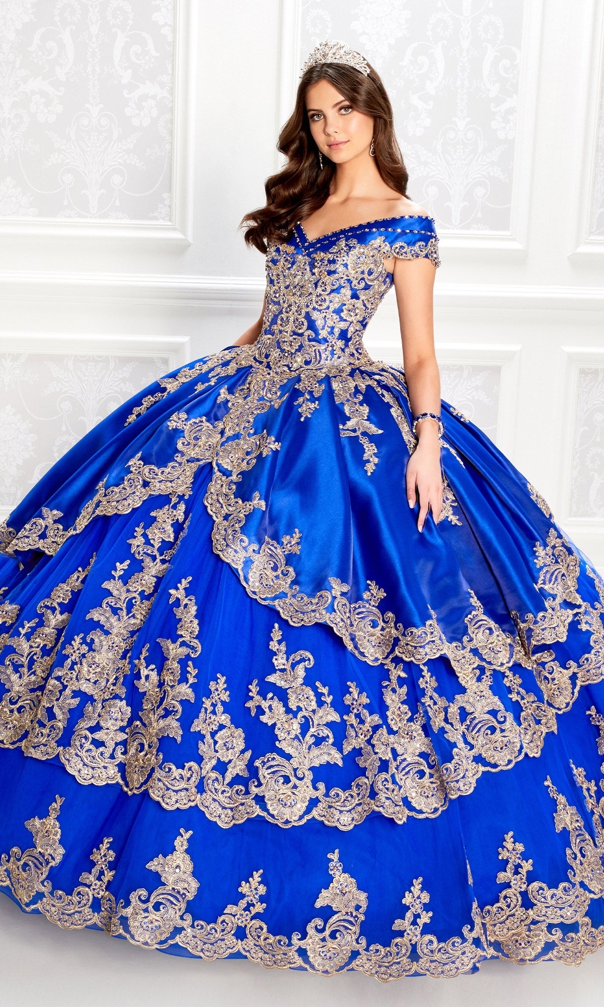 A young woman in the Princesa PR22029 Satin Quinceanera Dress, featuring off-the-shoulder blue satin with gold embroidery and a tiara, stands before a white decorative wall.